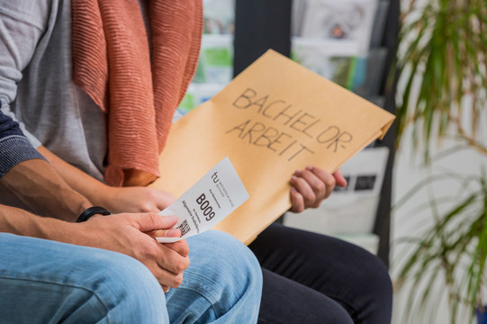 Close-up of two people sitting nect to each other. One is holding an envelope, the other a small piece of paper with a number printed on it