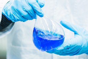 Two hands in blue rubber gloves hold a volumetric flask filled with blue liquid.